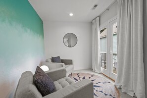 A modern living room with a grey sofa set, a round mirror on the wall, and a view of the outdoors through a sliding glass door. - Walk on Water: Soundside, Community Pool and Clubhouse Access (Kill Devil Hills)