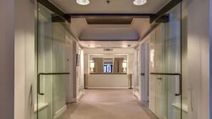 A modern hotel corridor with glass doors, a central light fixture, and a view into a room with a window and a door at the end.