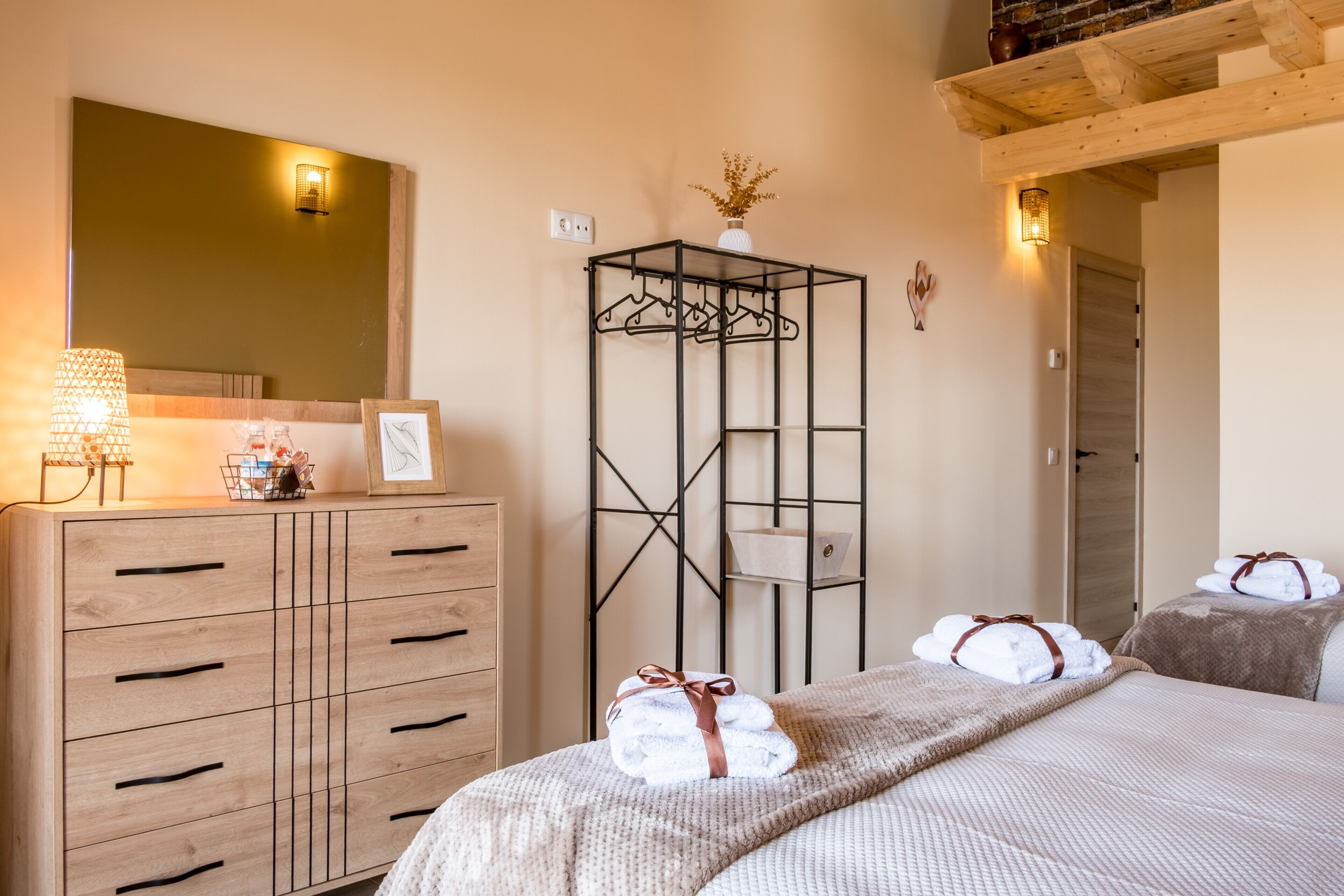 Habitación de hotel con cómoda de madera, perchero metálico, cama con toallas y espejo con apliques de pared.
