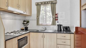 A compact kitchen with a gas stove, microwave, sink, and a window with curtains. - 2 Bedroom Budget Villa (Metung)