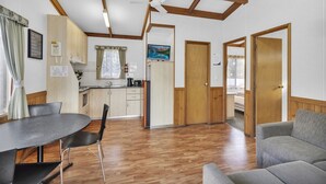 A spacious living area with a dining table, chairs, and a sofa. There is a kitchen area with wooden cabinets and a window. - 2 Bedroom Budget Villa (Metung)