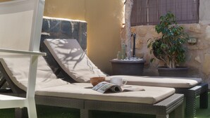 Un área de descanso al aire libre iluminada por el sol, con una silla blanca, un sillón acolchado, una planta en maceta y un muro de piedra.