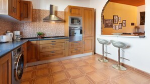 Una cocina con armarios de madera, lavadora, microondas y pileta. Hay dos taburetes de bar y un salpicadero de azulejos.