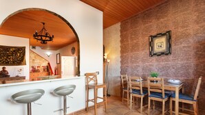 Un comedor con una mesa y sillas de madera, una barra con taburetes, una araña y una pared de piedra con un cuadro enmarcado.