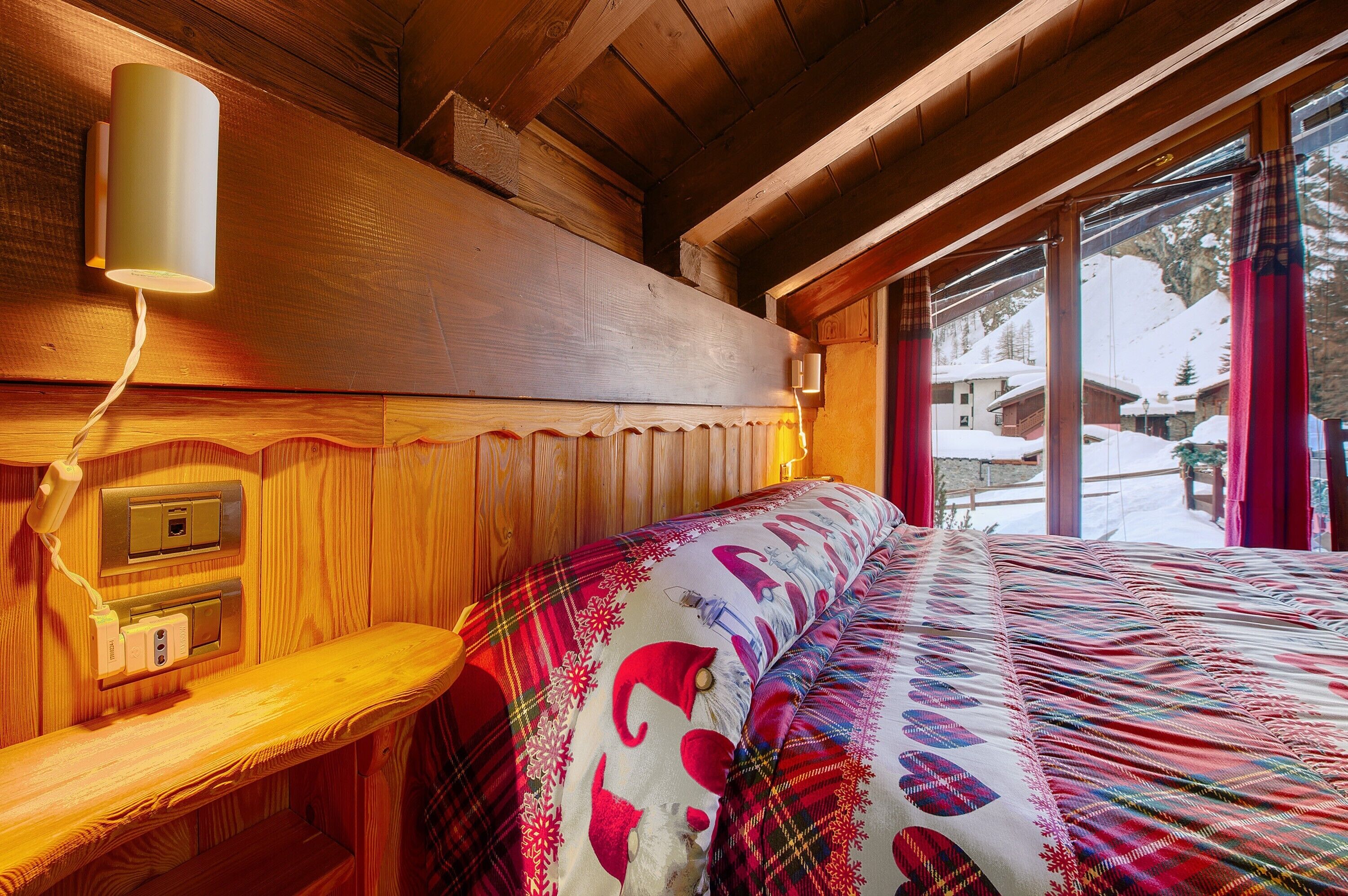 A wooden cabin interior with a bed, a wall-mounted lamp, and a window with a view of a snowy landscape.