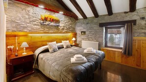 Un dormitorio con pared de piedra, vigas de madera en el techo, una cama con cubrecama gris, una mesita de noche con lámpara y una ventana con cortinas.