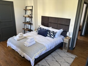 A neatly made bed with towels, a bedside table with a lamp, and a wall-mounted shelf with a tissue box and a decorative item. - Cosy Apartment in Eymet - Air Conditioned for 4 people - Trois Cles (Eymet)