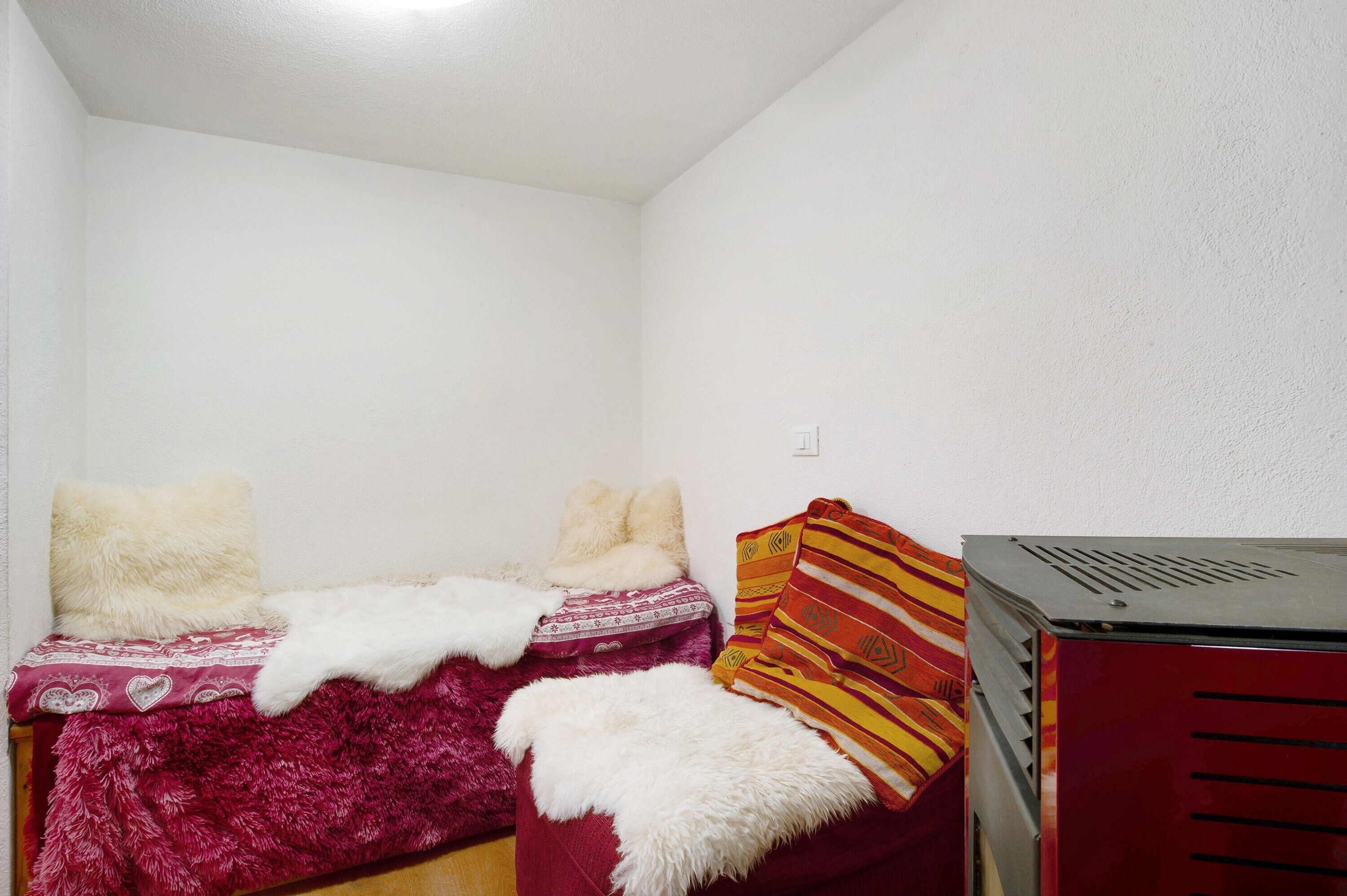 Een kleine kamer met een enkel bed, een rood-wit gebedde deken, zachte witte kussens en een rode verwarming.