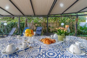 Un espace extérieur abrité, aménagé pour prendre les repas, avec une table dressée pour le petit-déjeuner, comprenant des croissants, des fruits et une plante en pot.