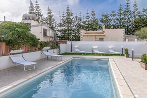 Une piscine entourée de chaises longues, d’un mur blanc et d’arbres.