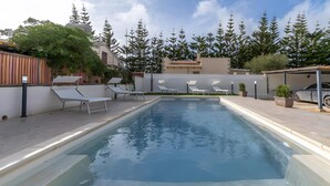 Ein rechteckiger Pool mit weißen Liegestühlen, einer überdachten Sitzgruppe und einer Baumreihe im Hintergrund.