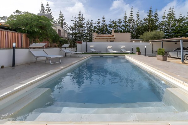 Une piscine rectangulaire bordée de chaises longues blanches, un espace de détente couvert et une rangée d’arbres en arrière-plan.