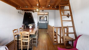 A wooden dining table with chairs, a fireplace, a kitchen in the background, and a wooden ladder. -  Le Carlit – Apartment in Les Angles with mountain view, cosy and functional (Les Angles)