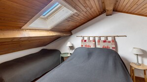 A small attic room with wooden beams, two beds, a skylight, and a small table with lamps. -  Le Carlit – Apartment in Les Angles with mountain view, cosy and functional (Les Angles)