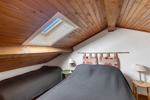 Une petite chambre mansardée avec des poutres en bois, deux lits, une lucarne et une petite table avec des lampes.