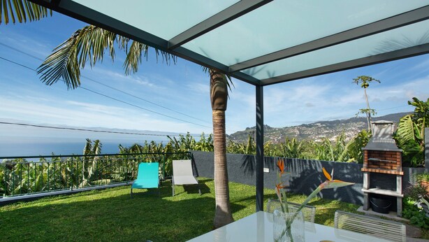 A patio with a glass enclosure, a white table, a vase with flowers, a grill, and a view of the ocean and hills.