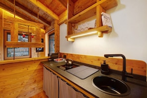 Interior de uma cabana de madeira com área de cozinha, incluindo pia, chaleira e prateleiras de madeira.