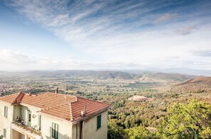 A building with a red-tiled roof, a balcony, and a view of a valley with hills and a town in the distance.