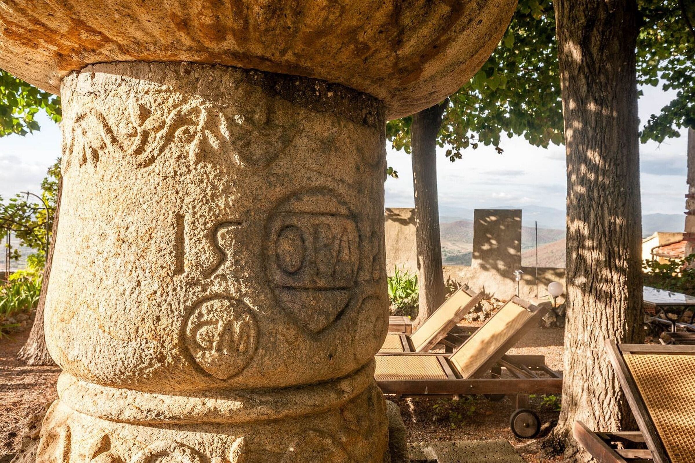 A stone pillar with carvings, a wooden lounge chair, and a view of the landscape.
