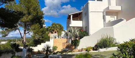A modern white building with a stone base, surrounded by lush greenery and trees.