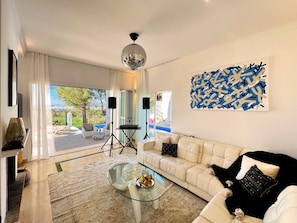 A modern living room with a glass coffee table, white sofa, and a view of a pool and greenery. - Holiday Home 'Sunset Bay' with Sea View, Wi-Fi and Air Conditioning (Illes Balears)