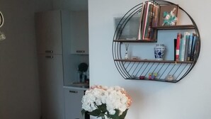 Un comedor con una mesa blanca, sillas con estampado floral negro, un jarrón de flores blancas, un pequeño pizarrón y una estantería metálica con libros y objetos decorativos.
