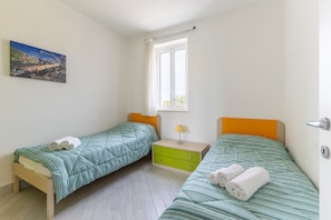 A bedroom with two beds, a window, a bedside table with a lamp, and a painting on the wall.