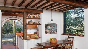 A rustic kitchen with wooden beams, a dining table with benches, a window with a view of trees, and a shelf with kitchenware. - Holiday Home 'Chianti Loft' with Mountain View, Wi-Fi and Air Conditioning (Greve in Chianti)