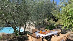 A round table with wicker chairs, a fruit bowl, and a tray with drinks, set against a backdrop of olive trees and a pool. - Holiday Home 'Chianti Loft' with Mountain View, Wi-Fi and Air Conditioning (Greve in Chianti)