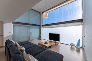 A modern living room with a large TV, a sofa, a coffee table, and a view through large windows.