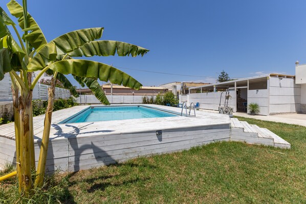 A swimming pool with a wooden deck, surrounded by a grassy area and a banana tree. - Apartment 'Stella Marina' with Shared Pool, Wi-Fi and Air Conditioning (Pachino)