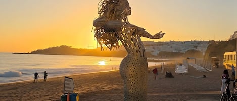 A sculpture of a mermaid on a beach with a sunset in the background.