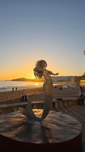 A sculpture of a mermaid on a beach with a sunset in the background.