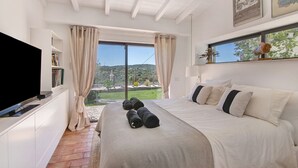 Een slaapkamer met een bed, een televisie, een raam met gordijnen en uitzicht op de omgeving.