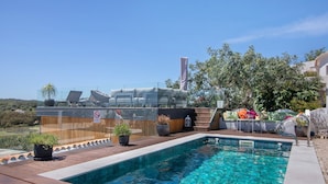 Una piscina con sillas de descanso y plantas en macetas, rodeada por una terraza de madera y una valla de vidrio.