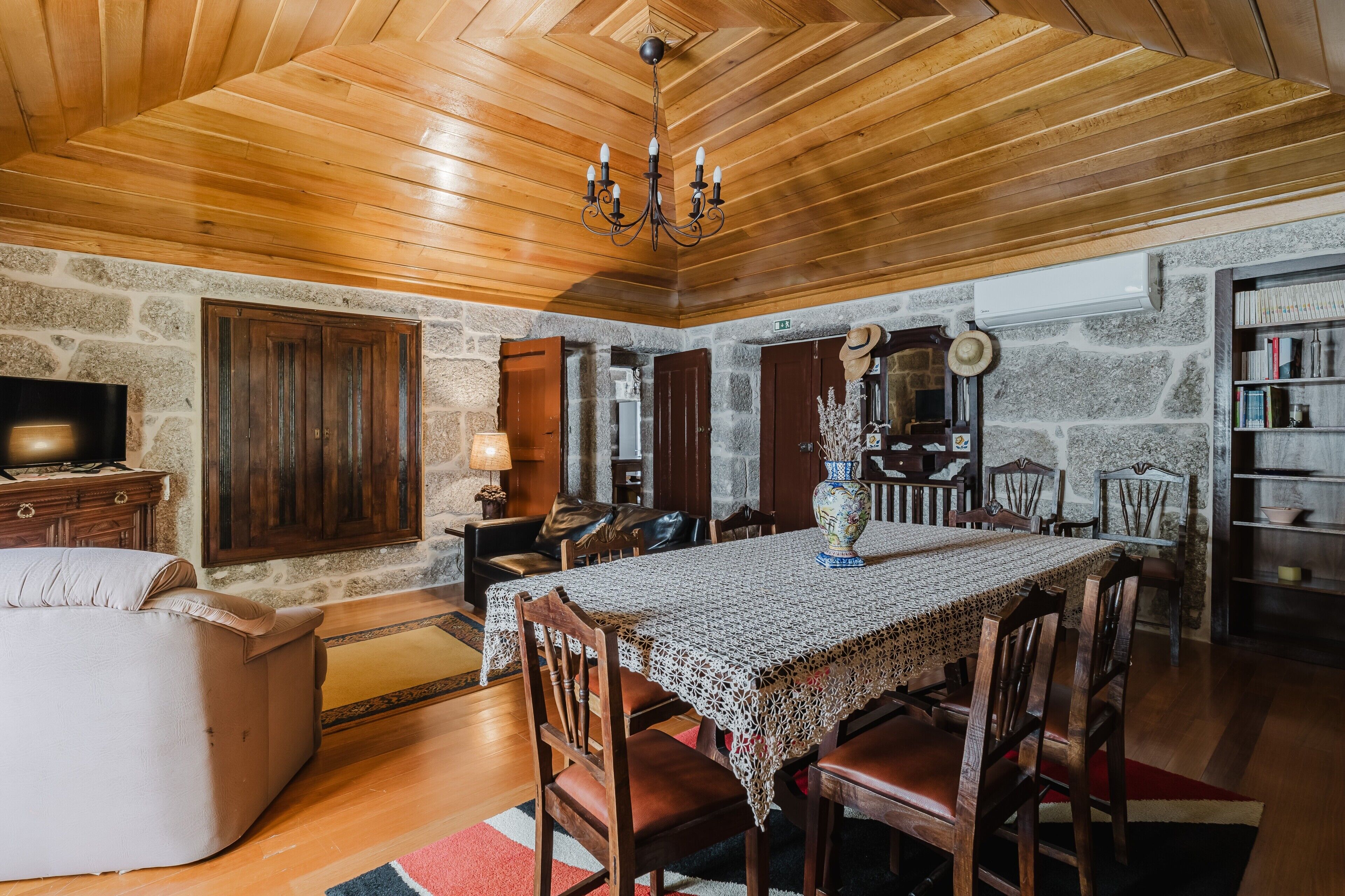 Un comedor con techo de madera, paredes de piedra, una araña, una mesa con mantel de encaje, sillas de madera y una estantería.