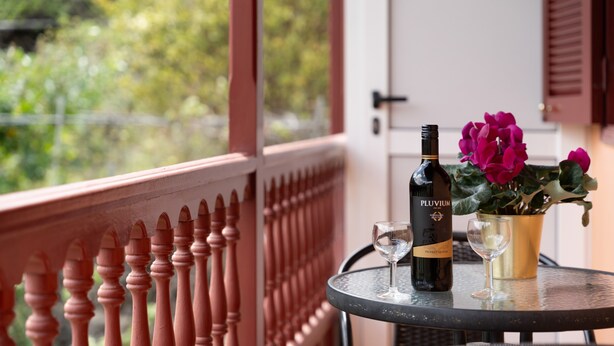 Een balkon met een houten hekwerk, een tafel met een fles en een glas, en een bloempot met roze bloemen.