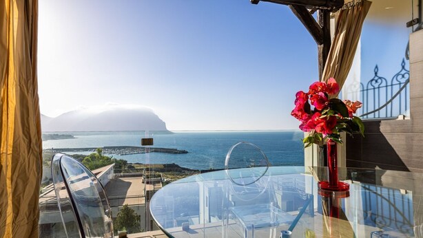 A glass table with a red vase of flowers, overlooking a coastal view with a clear sky and distant landmass.