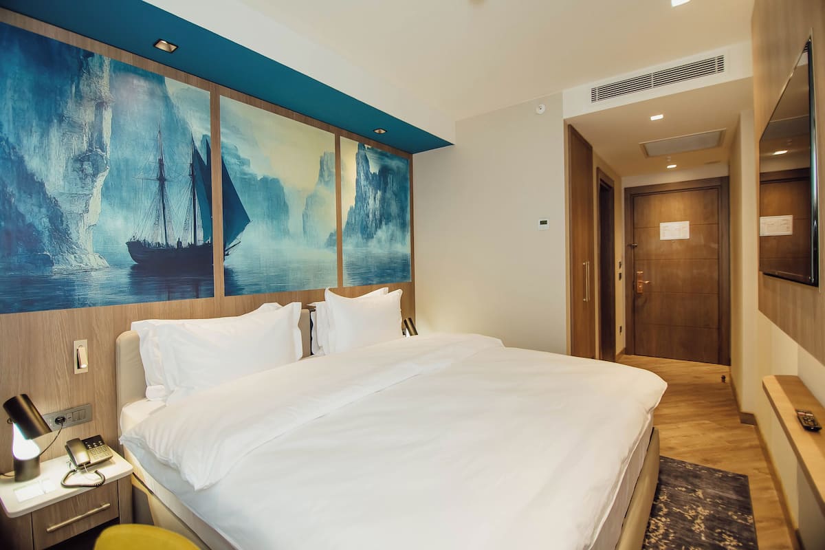 A hotel room with a large bed, a nightstand with a lamp and phone, a painting of a sailboat, and a wooden door.