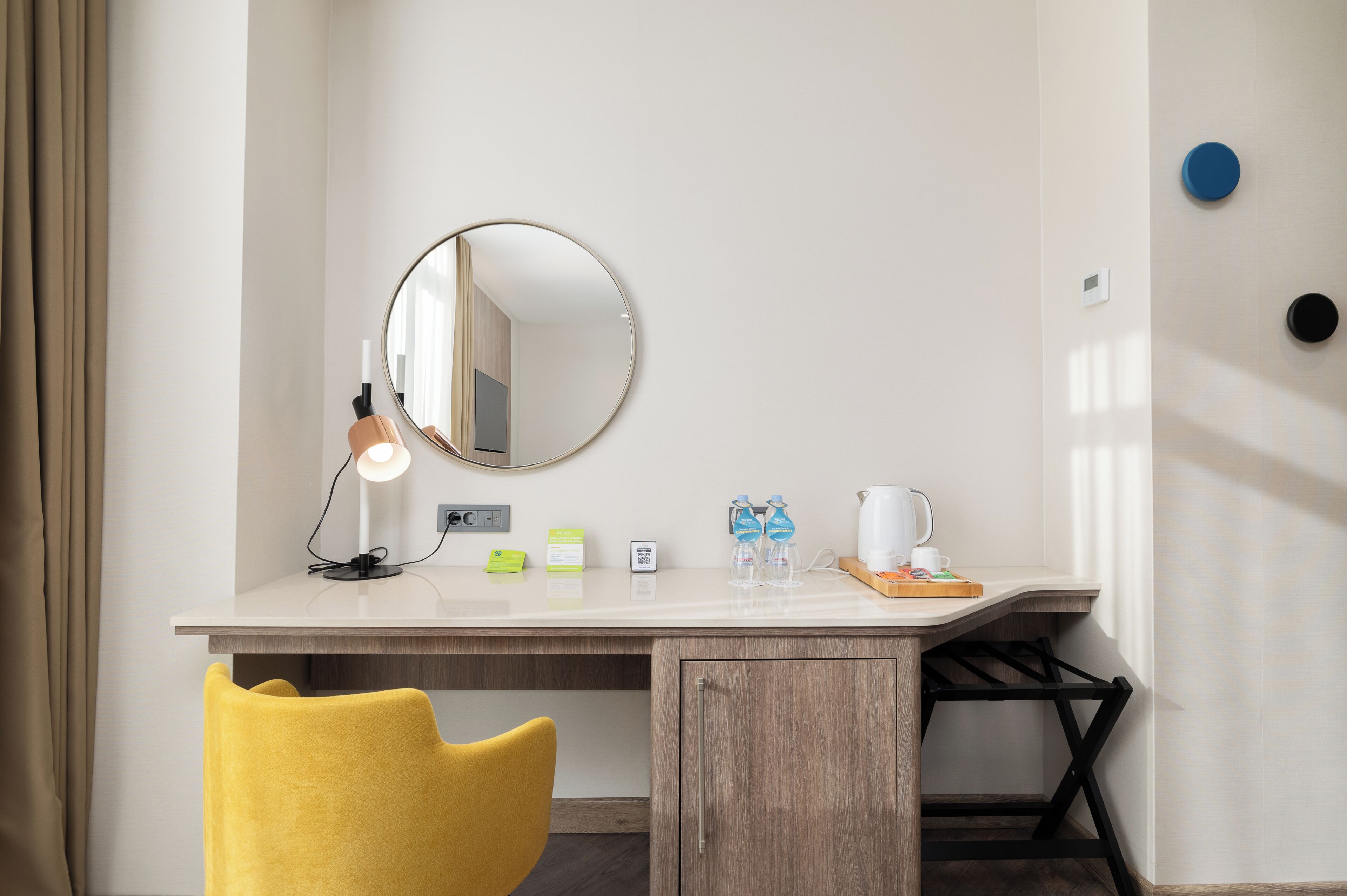 A hotel room with a desk, a yellow chair, a round mirror, a lamp, a coffee maker, and a small shelf with toiletries.