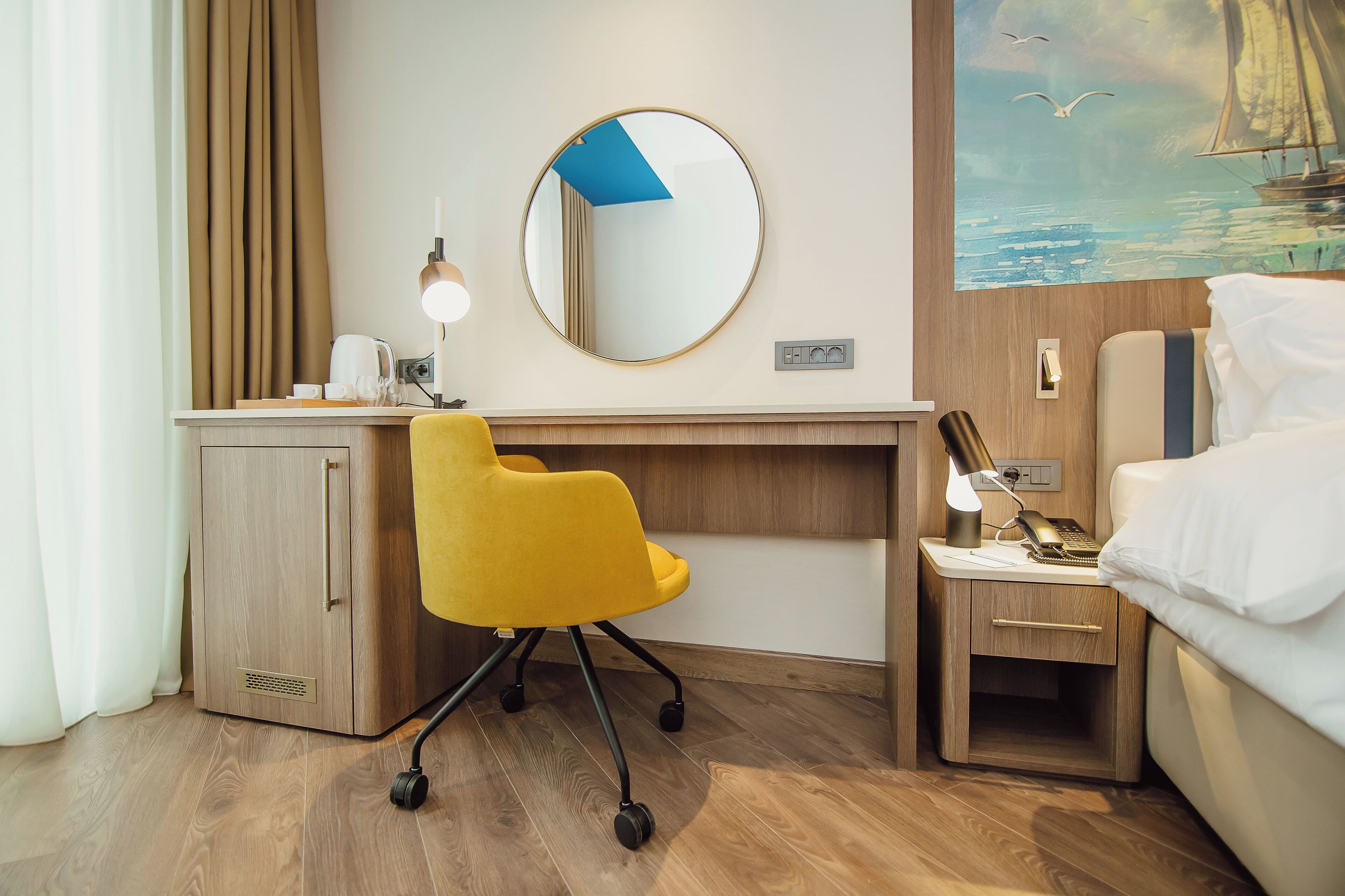 Hotelkamer met een houten bureau, een gele stoel, een bed, een nachtkastje, een lamp en een schilderij van een zeilboot aan de muur.