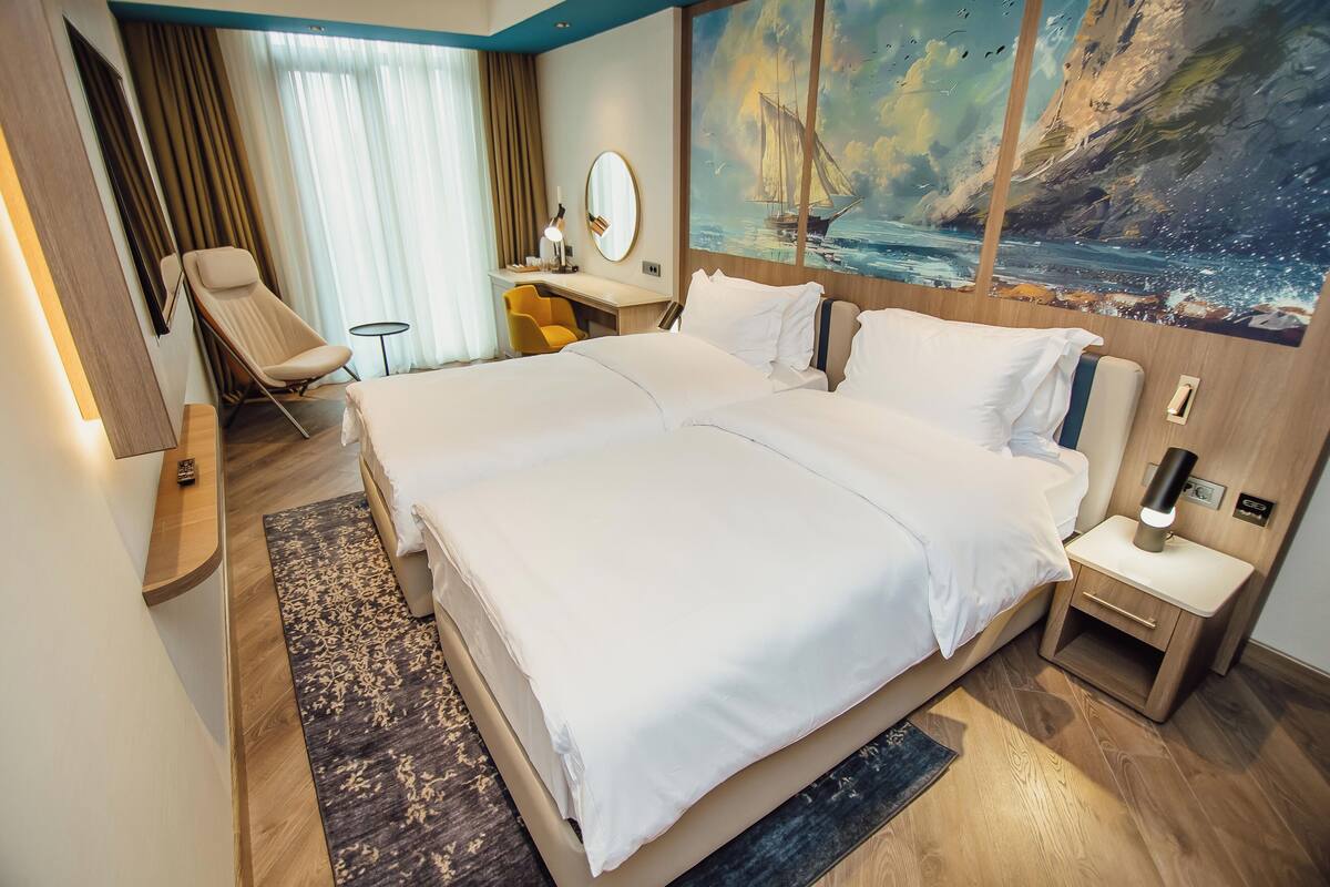 Una camera d'albergo con un letto grande, una scrivania con una sedia, un tavolino, uno specchio e un quadro raffigurante una barca a vela appeso al muro.