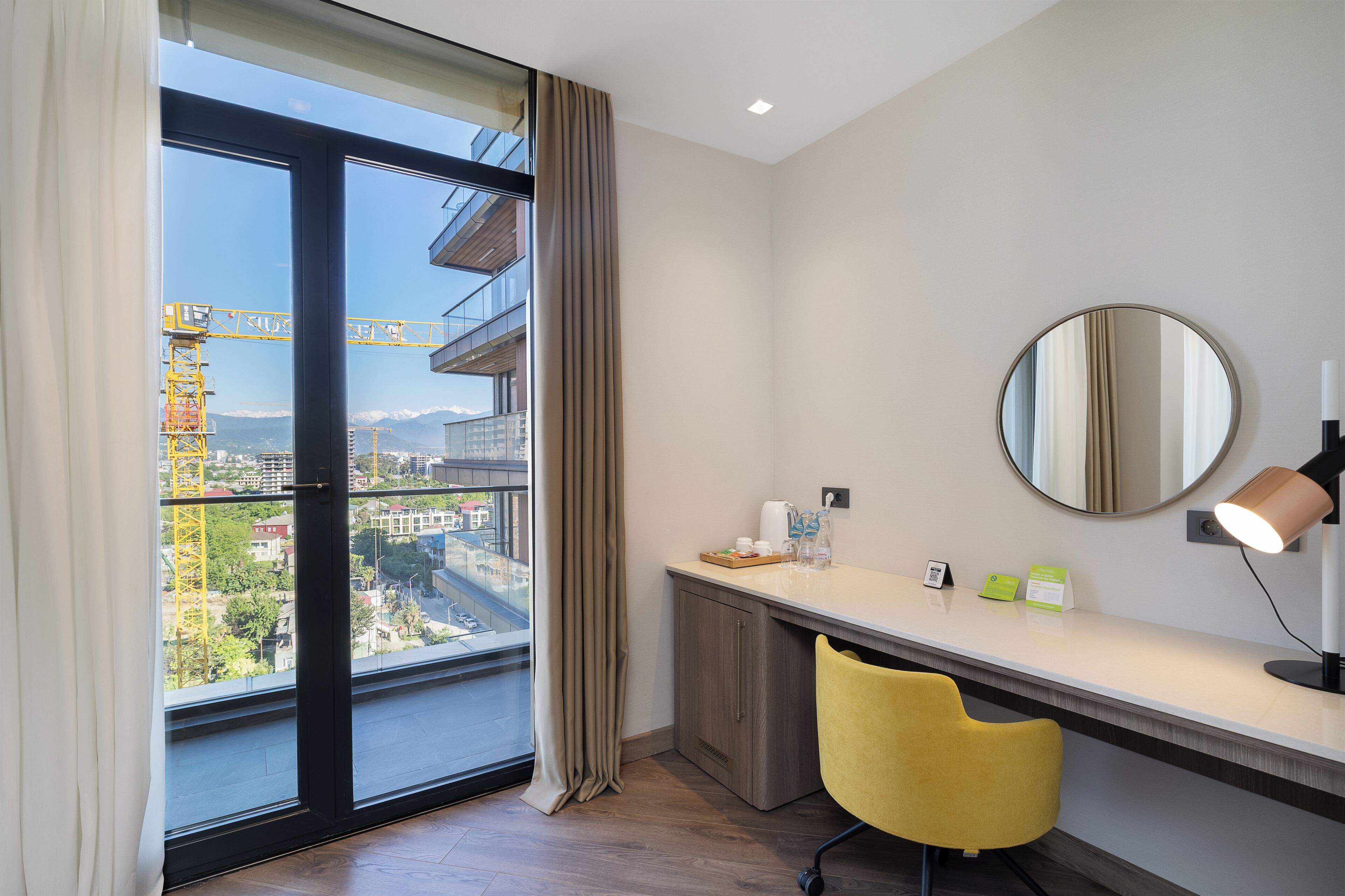 A modern hotel room with a large window, a desk with a chair, a mirror, and a lamp.
