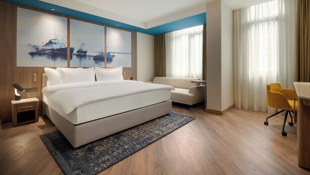 A modern hotel room with a large bed, a desk, a chair, and a painting of boats on the wall. - La Quinta by Wyndham Batumi (Batumi)