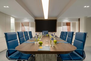 A conference room with a long table, blue chairs, and a large screen at the front.