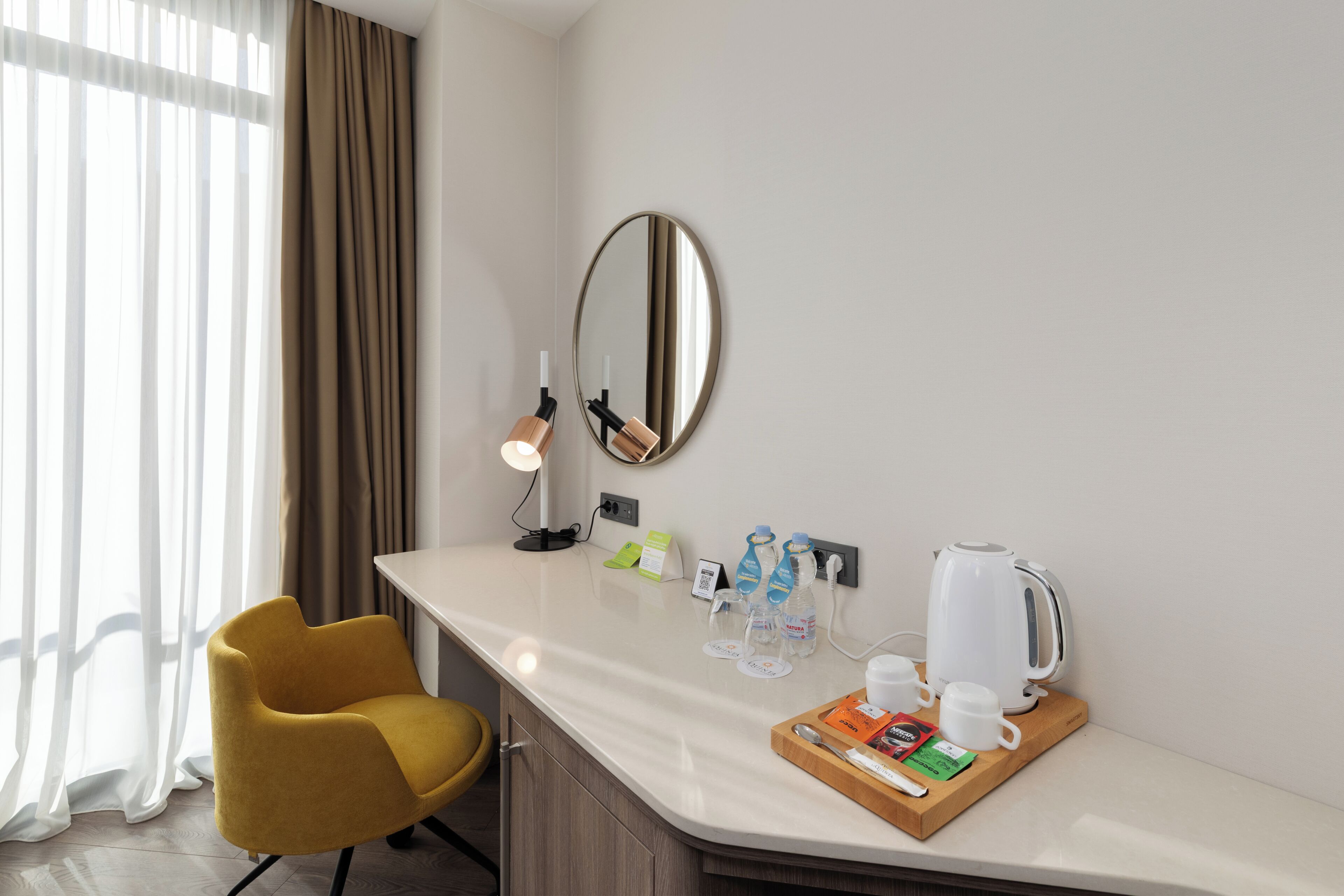 A hotel room with a white countertop, a yellow chair, a mirror, a lamp, a kettle, and a tray with cups and a spoon.