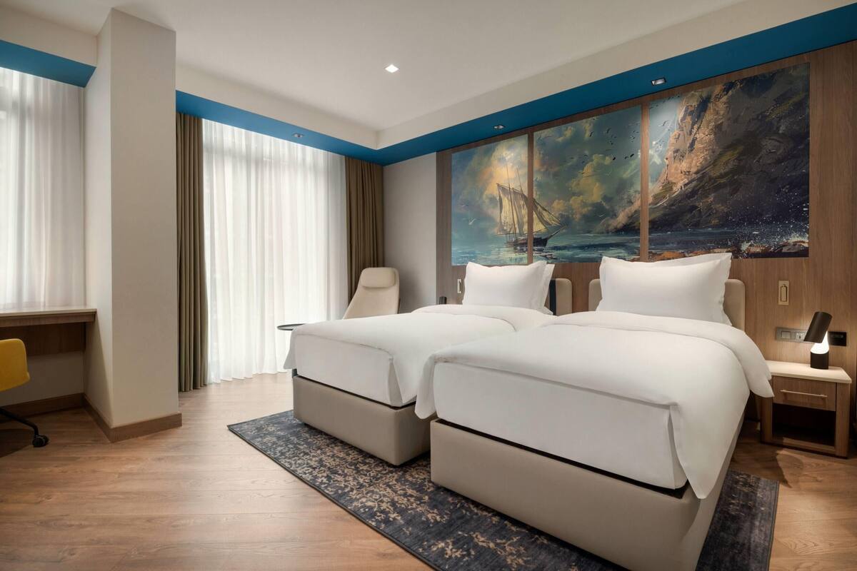Camera d'albergo con due letti, una scrivania, una sedia, un comodino con lampada e un quadro di grandi dimensioni raffigurante una veduta costiera.