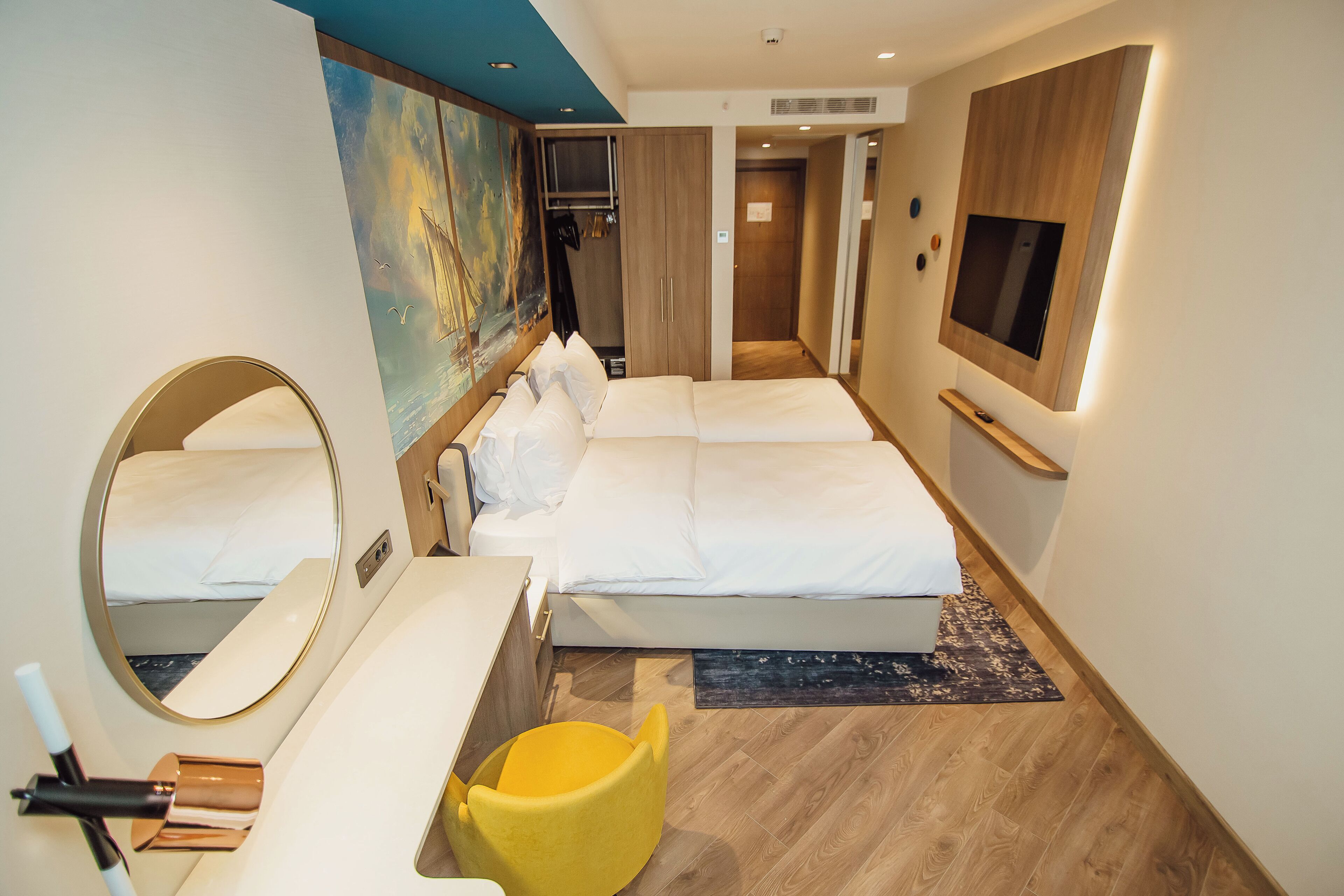 A hotel room with a large bed, a bedside table, a mirror, a yellow chair, and a TV mounted on the wall.