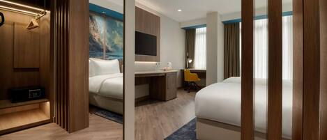 A modern hotel room with a wooden wardrobe, a desk with a chair, a bed, and a large painting on the wall.