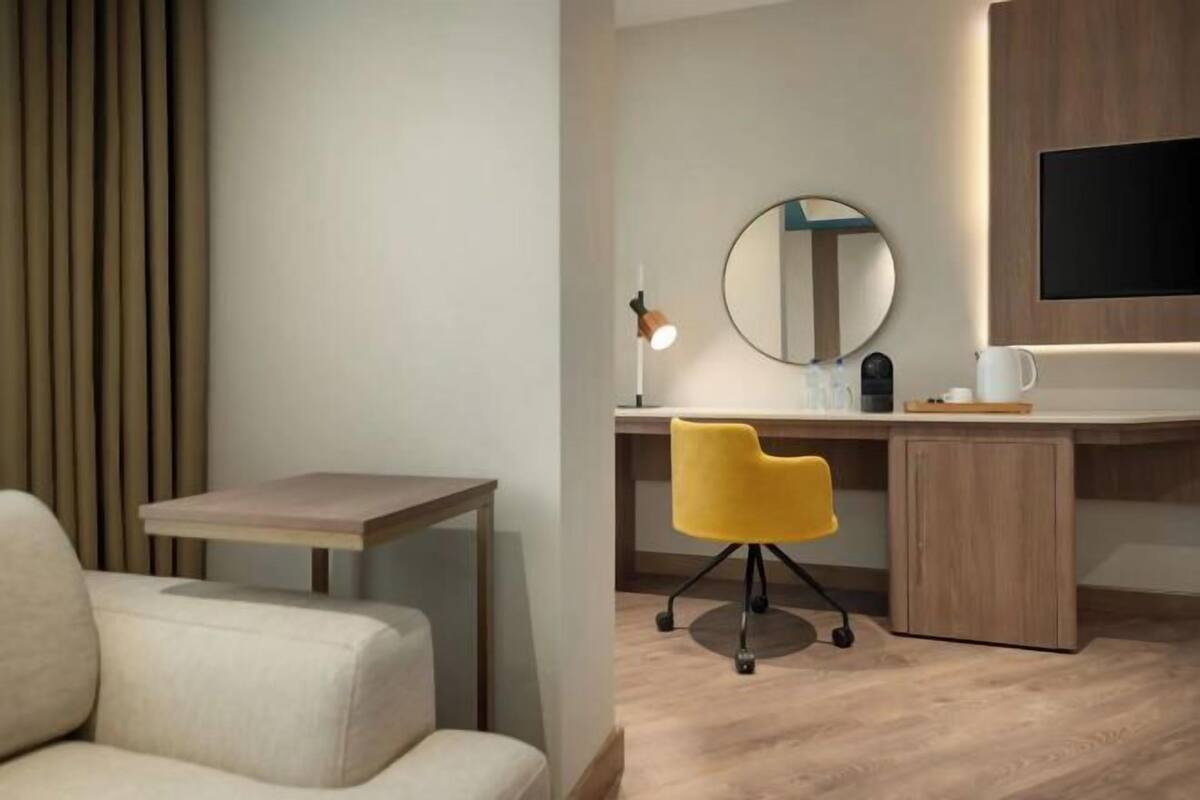 Una moderna camera d'hotel con una scrivania in legno, una sedia gialla, uno specchio rotondo, una TV a schermo piatto e un divano.